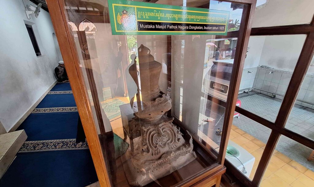 20260508_121235 - Mabur.co Glass-front display case housing a carved stone sculpture on a wooden pedestal, with a green-yellow sign above; a man sits praying on a blue carpeted hallway to the left.