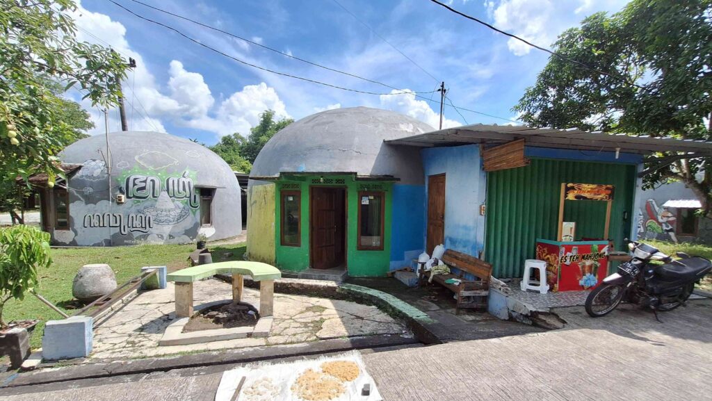 20260525_112418 - Mabur.co Two gray domed houses with colorful green and blue accents sit along a sunny street, with a small outdoor seating area and a parked motorcycle nearby.