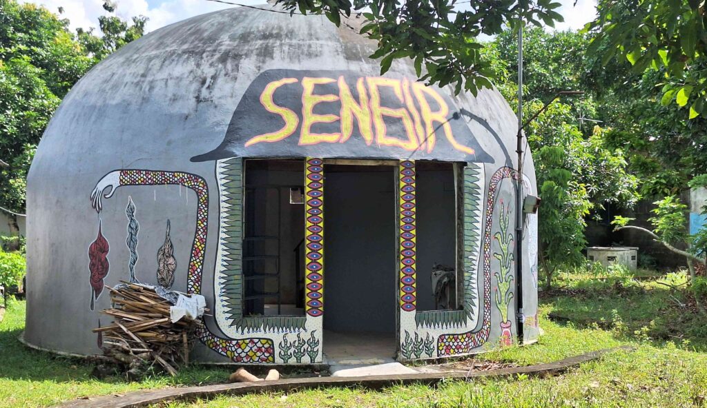 20260525_112520 - Mabur.co Round concrete hut with a painted mural reading 'SENHOR' on the front, framed by yellow and pink doorway columns and decorative patterns, set among trees and greenery.