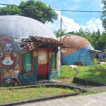Colorful dome-shaped buildings with large cartoon mural outside, red doors, and a tiled roof, set in a sunny tropical yard.