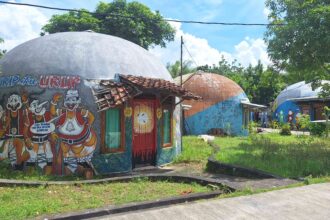 Colorful dome-shaped buildings with large cartoon mural outside, red doors, and a tiled roof, set in a sunny tropical yard.