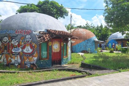 Colorful dome-shaped buildings with large cartoon mural outside, red doors, and a tiled roof, set in a sunny tropical yard.