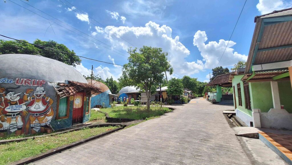 20260525_112551 - Mabur.co Sunny street in a small neighborhood with a domed building painted with cartoon characters on the left and green houses on the right.