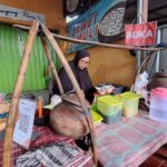 Woman in a black hijab cooking at a street stall with large wooden cooking pot and colorful plastic containers; green shuttered shop in background.