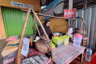 Woman in a black hijab cooking at a street stall with large wooden cooking pot and colorful plastic containers; green shuttered shop in background.