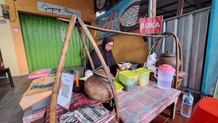 Woman in a black hijab cooking at a street stall with large wooden cooking pot and colorful plastic containers; green shuttered shop in background.