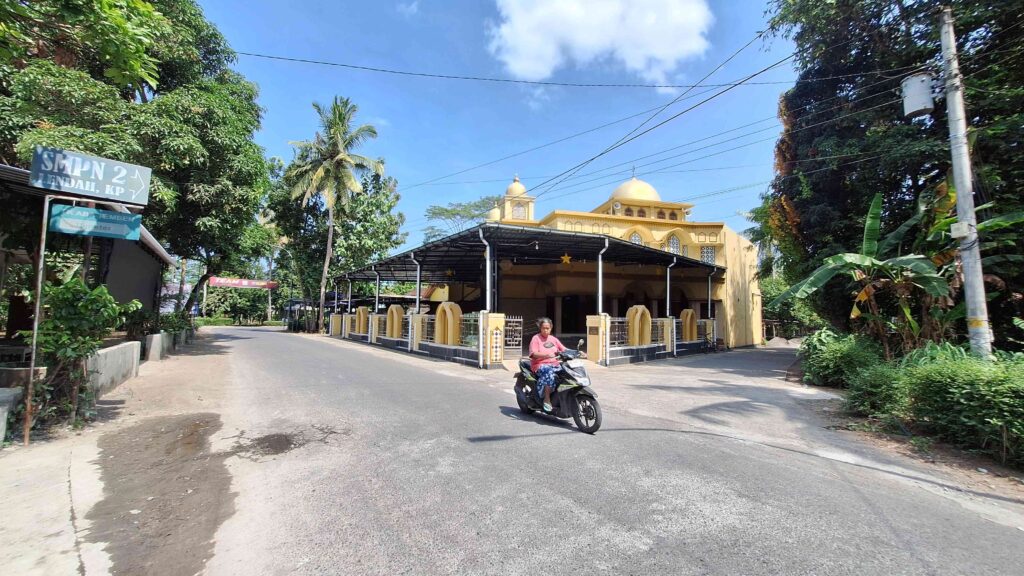 Yellow mosque with domed roof and arches on a sunny street; a woman on a scooter rides toward the camera.