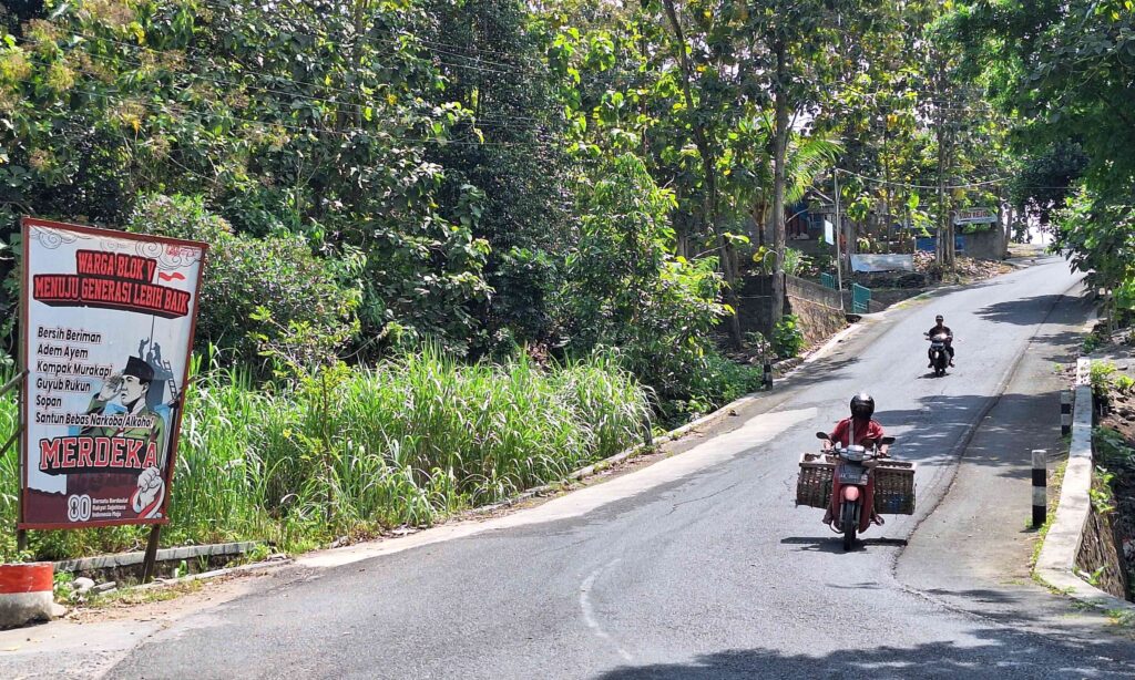 Rural street scene with motorcycles riding on a paved road, dense trees on both sides and a large signboard to the left showing 'MERDEKA' and Indonesian slogans.