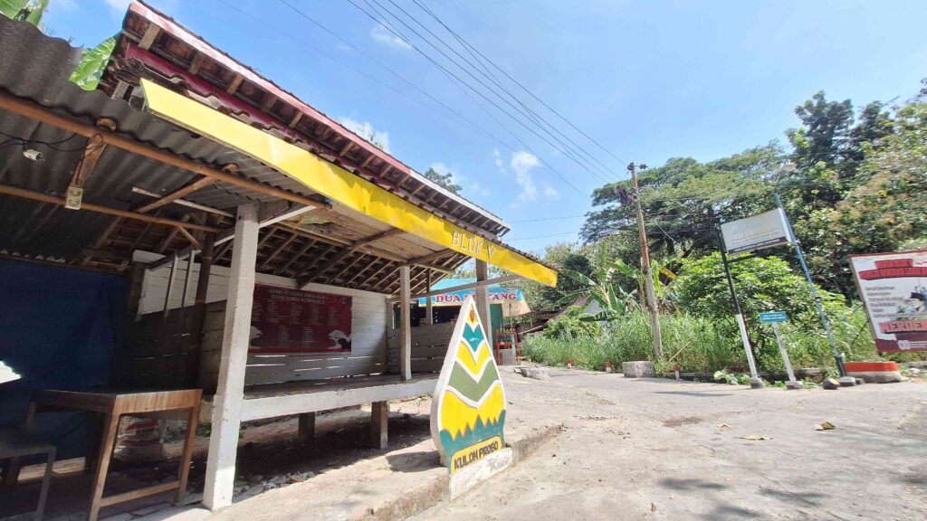 Open-front market stall with a yellow beam labeled 'BLOK V' and a large colorful chevron road sign in the foreground under a clear blue sky.