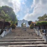 Stone staircase leading to a white temple archway at the top, flanked by trees and low walls under a blue sky with clouds.