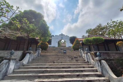Stone staircase leading to a white temple archway at the top, flanked by trees and low walls under a blue sky with clouds.