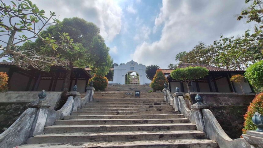 20260603_120454 - Mabur.co Stone staircase leading to a white temple archway at the top, flanked by trees and low walls under a blue sky with clouds.