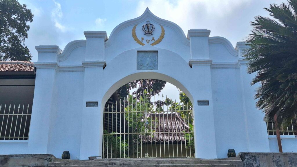 20260603_120531 - Mabur.co White arched gateway with a gold laurel wreath, crown emblem, and a gated entrance set in a light blue wall; tropical plants nearby