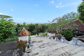Stone steps and decorative balustrades descend into a lush courtyard with temple-style, tiled-roof structures and a blue sky above.