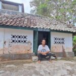 A man sits barefoot on the steps outside a small white-washed building with a weathered tiled roof in a rural setting.