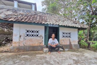 A man sits barefoot on the steps outside a small white-washed building with a weathered tiled roof in a rural setting.
