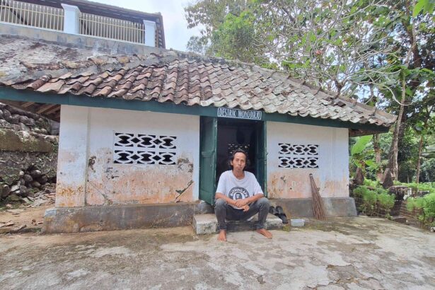 A man sits barefoot on the steps outside a small white-washed building with a weathered tiled roof in a rural setting.