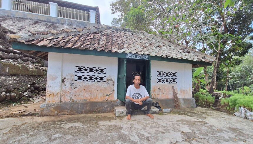 20260603_121641 - Mabur.co A man sits barefoot on the steps outside a small white-washed building with a weathered tiled roof in a rural setting.