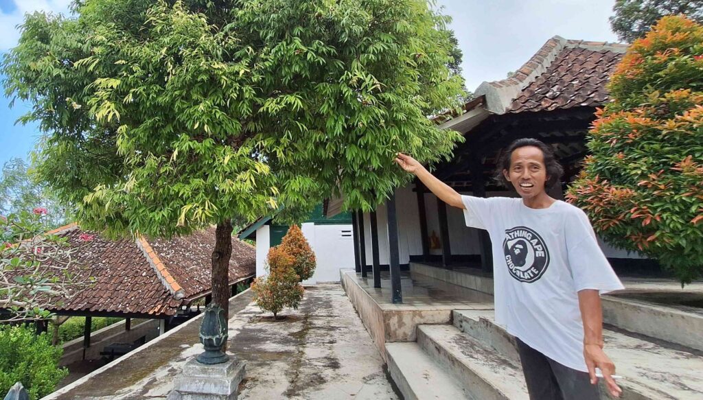 20260603_122917 - Mabur.co Man in a white T-shirt standing on a concrete path, gesturing toward a large leafy tree beside a building with a tiled roof