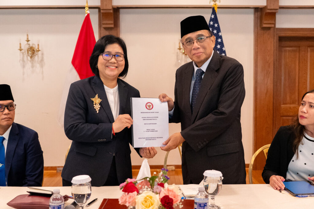 Two professionals exchange a formal document at a ceremony, with Indonesian and US flags in the background and others seated nearby.