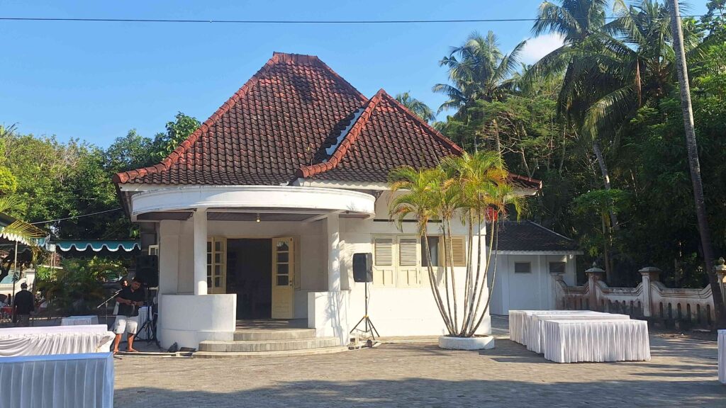 Small white pavilion with a red tiled roof in a tropical setting, surrounded by palm trees and rows of white-covered tables for an outdoor event.