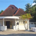 Small white pavilion with a red tiled roof in a tropical setting, surrounded by palm trees and rows of white-covered tables for an outdoor event.