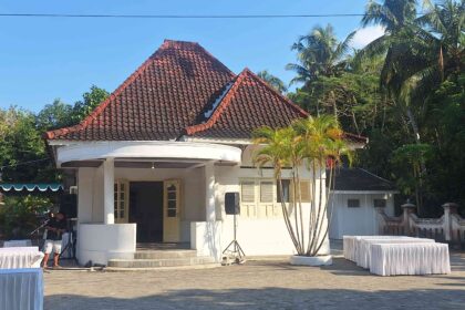 Small white pavilion with a red tiled roof in a tropical setting, surrounded by palm trees and rows of white-covered tables for an outdoor event.