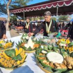 Men in black traditional attire and gold sashes stand around a table of elaborate fruit baskets under a decorated canopy at an outdoor ceremony.