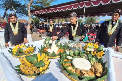 Men in black traditional attire and gold sashes stand around a table of elaborate fruit baskets under a decorated canopy at an outdoor ceremony.