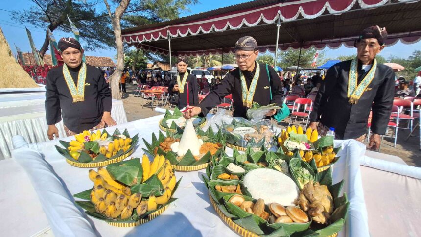 Men in black traditional attire and gold sashes stand around a table of elaborate fruit baskets under a decorated canopy at an outdoor ceremony.