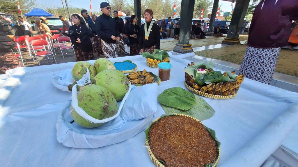 Outdoor cultural ceremony with people in black traditional clothing; a long table displays baskets of fruit, leaves, and spices under a pavilion.