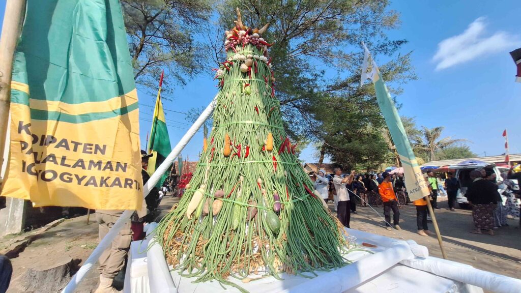 Tall cone-shaped centerpiece made of green vegetables and peppers at an outdoor festival, with banners and spectators nearby.