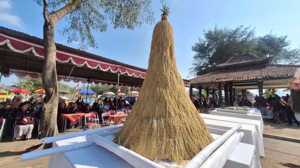 Crowd seated at an outdoor festival under a red-and-white canopy, with a tall straw sculpture centerpiece in the foreground.