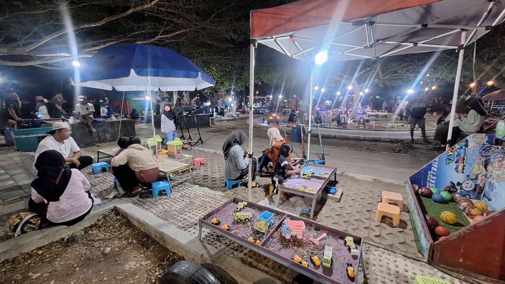 20260627_195737 - Mabur.co Night market or festival scene at night with families under tents, children playing with toy booths and small plastic chairs visible on the ground.