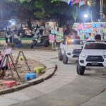 Children draw on easels at a lively outdoor night market while parents watch; small white toy cars drive along a paved street nearby.