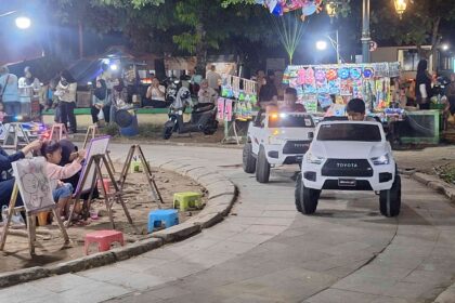 Children draw on easels at a lively outdoor night market while parents watch; small white toy cars drive along a paved street nearby.