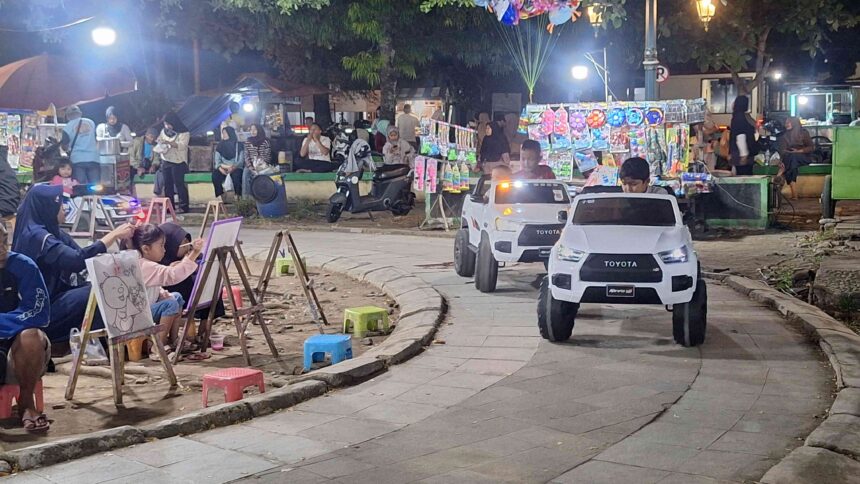 20260627_195853 - Mabur.co Children draw on easels at a lively outdoor night market while parents watch; small white toy cars drive along a paved street nearby.