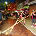 Two musicians in traditional outfits play long wooden alphorns on a brick-paved street at night during a festive evening crowd.