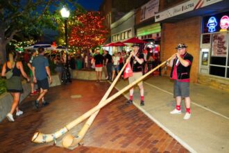 Two musicians in traditional outfits play long wooden alphorns on a brick-paved street at night during a festive evening crowd.
