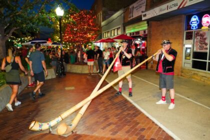 Two musicians in traditional outfits play long wooden alphorns on a brick-paved street at night during a festive evening crowd.