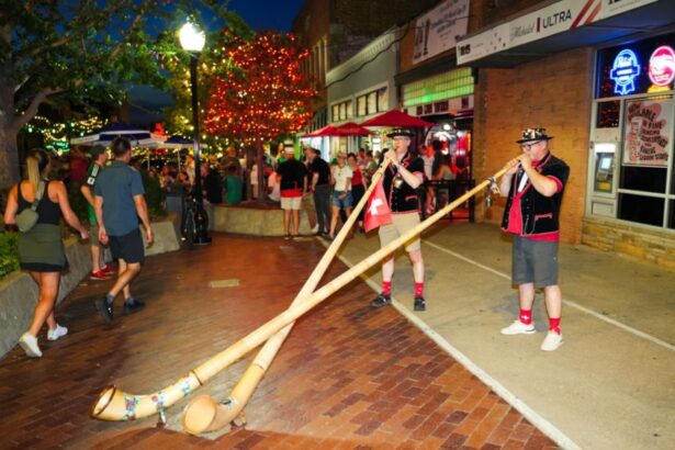Two musicians in traditional outfits play long wooden alphorns on a brick-paved street at night during a festive evening crowd.