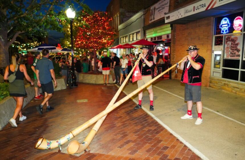 Two musicians in traditional outfits play long wooden alphorns on a brick-paved street at night during a festive evening crowd.