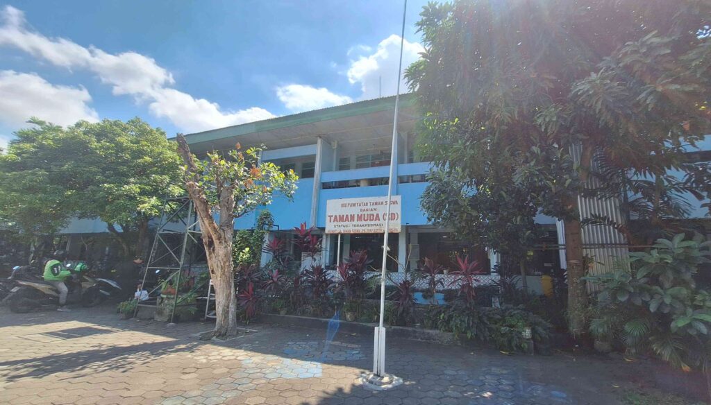 Blue two-story building with a sign reading 'TAMAN MUDA' amid trees in a sunny courtyard near a scooter.