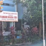 Signboard in Indonesian reading 'Taman Muda (SD)' in front of a blue building, surrounded by trees and red-leaf plants.