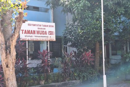 Signboard in Indonesian reading 'Taman Muda (SD)' in front of a blue building, surrounded by trees and red-leaf plants.
