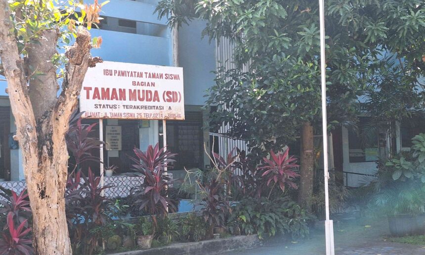 Signboard in Indonesian reading 'Taman Muda (SD)' in front of a blue building, surrounded by trees and red-leaf plants.