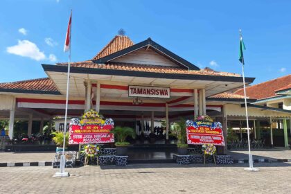 Stage area of a campus with red-and-white drapes, two flagpoles (Indonesia flags) and a banner reading TAMANSISWA; festive flower arrangements surround the entrance.