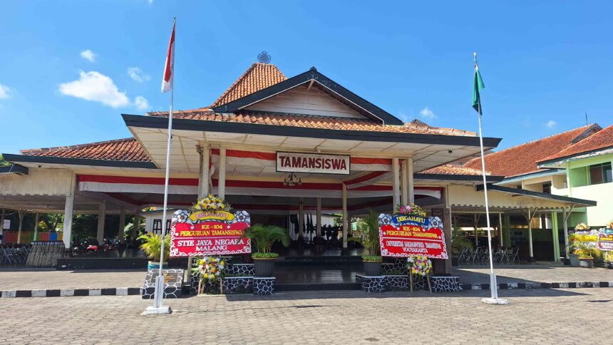 Stage area of a campus with red-and-white drapes, two flagpoles (Indonesia flags) and a banner reading TAMANSISWA; festive flower arrangements surround the entrance.