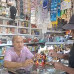 A shop cashier in a pink shirt hands an item to a customer across a glass counter, with shelves full of products behind them.