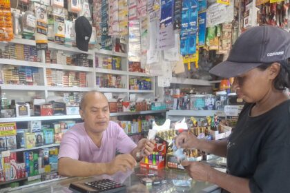 A shop cashier in a pink shirt hands an item to a customer across a glass counter, with shelves full of products behind them.
